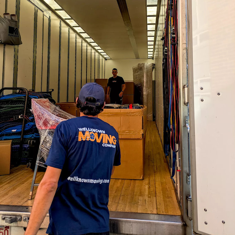 Two movers loading a large moving truck with various items including boxes and furniture. One mover is inside the truck organizing items while the other, wearing a blue company shirt, is standing at the truck's entrance overseeing the process.
